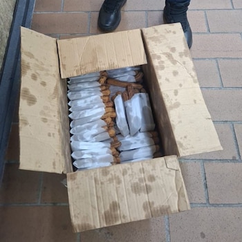 Una caja de cartón abierta con churros envueltos en papel sobre un suelo de baldosas y los pies de una persona con botas