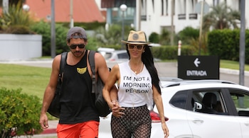 Pareja camina de la mano. Él con gorra, gafas de sol y camiseta negra. Ella con sombrero, gafas y camiseta blanca con mensaje. Fondo de vegetación y edificios.