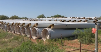 Gran cantidad de palas de turbinas eólicas blancas apiladas en filas largas sobre tierra seca y hierba bajo un cielo azul claro