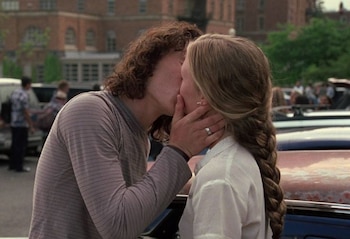 Una mujer con cabello rubio trenzado y un hombre con cabello rizado oscuro se besan apasionadamente, la mujer tiene un anillo en el dedo, con un fondo borroso de un campus y coches