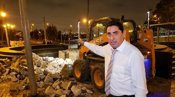 Carlos Álvarez, en camisa blanca y corbata, de pie junto a una máquina retroexcavadora naranja y escombros, apuntando en un paisaje nocturno urbano.