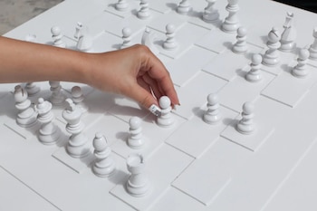 Mano con uñas pintadas mueve un peón blanco sobre un tablero de ajedrez blanco. Varias piezas blancas de ajedrez están dispuestas