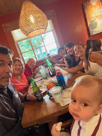 Un grupo de adultos y un bebé sonríen sentados a una mesa de madera con botellas y vasos en un restaurante