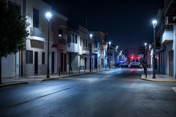 Calle urbana de noche iluminada por farolas con un coche patrulla y una ambulancia estacionados al fondo con luces encendidas.