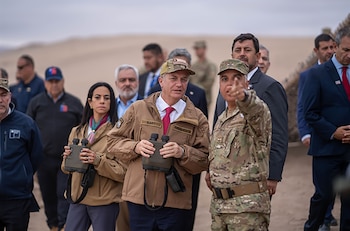Un grupo de hombres, incluyendo militares y funcionarios, se congrega al aire libre en un paisaje desértico, bajo una bandera chilena. Miran un caballete