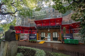 Edificio de ladrillos bajo restauración con andamios y una gran lona que cubre la fachada, frente a un busto de piedra, rodeado de frondosa vegetación y árboles