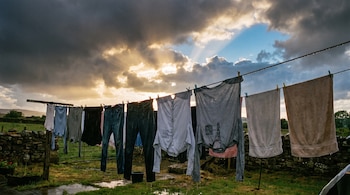 Prendas de ropa y toallas cuelgan de un tendedero con gotas. El suelo de césped está mojado con charcos. El cielo muestra nubes oscuras y sol.