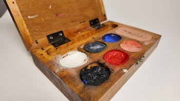 Caja de madera abierta con compartimentos de maquillaje teatral en tonos blanco, negro, rojo, azul, melocotón y piel, sobre una superficie blanca