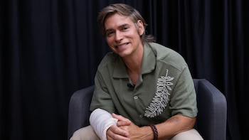 Carlos Baute sentado en silla, camisa verde con bordados, pantalón beige, brazo izquierdo vendado. Pulsera marrón en muñeca derecha, micrófono en camisa, fondo negro