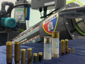 Dos militares en uniforme camuflado escuchan a un técnico con guantes de trabajo frente a estantes metálicos con pistolas en estuches