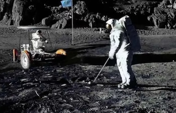 Astronauta en traje espacial blanco golpea una pelota de golf en la superficie lunar gris. Un vehículo lunar y una bandera azul se ven al fondo