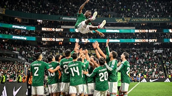 Un grupo de futbolistas con camiseta verde y pantalón blanco lanza al aire a otro jugador en un estadio con público.