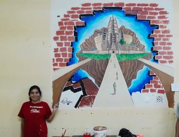 Mujer sonriente de cabello oscuro y camiseta roja, posando frente a un mural con una ciudad futurista flotante y una pared de ladrillos