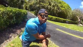 Hombre con gorra, gafas de sol y polo azul se arrodilla en el césped junto a una carretera con doble línea amarilla y densa vegetación