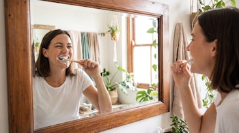 Mujer de cabello castaño se cepilla los dientes con cepillo de bambú, espuma en la boca, frente a espejo de marco de madera en un baño con plantas y toallas.