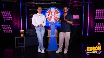 Dos hombres de pie en un estudio de televisión. Al centro, una ruleta azul y blanca. A los lados, luces rosas. Hay un cubo de palomitas en una mesa y un sofá negro