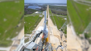Vista aérea de un cohete blanco y naranja en una plataforma de lanzamiento de hormigón, con una carretera recta y edificios en un paisaje verde y acuático