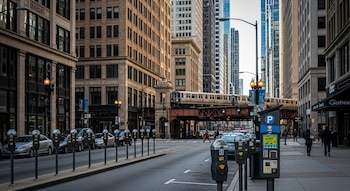 Vista de una calle urbana en Chicago con edificios altos, un tren elevado, múltiples parquímetros, coches aparcados y peatones caminando.