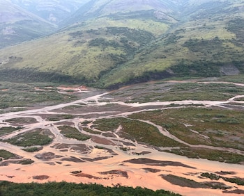 Vista aérea de un valle montañoso verde con un río trenzado de múltiples canales de color rojizo y grisáceo. La vegetación cubre las laderas y orillas