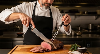 Un chef con barba y delantal negro corta carne asada rosada con un cuchillo y tenedor en una tabla de madera, con romero y sal cerca, en una cocina.