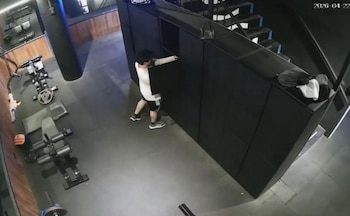 Vista aérea de un hombre en camiseta blanca y pantalones cortos negros abriendo un casillero oscuro en un gimnasio. Se ve equipamiento y el suelo de goma