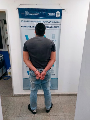Un hombre de espaldas, con camiseta gris y pantalón de jean, tiene las manos esposadas. Está de pie frente a un cartel de la Policía de Bahía Blanca