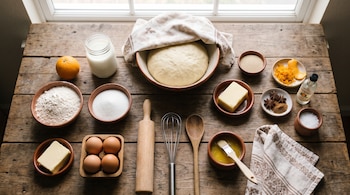 Recipientes con harina, huevos, mantequilla, azúcar, leche, ralladura de naranja, especias, masa fermentada y utensilios sobre una mesa de madera.