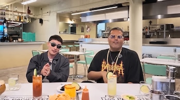 Dos hombres sentados en una mesa con micrófonos, bebidas y comida. En el fondo se ven mesas, sillas, un bar y la cocina del restaurante.