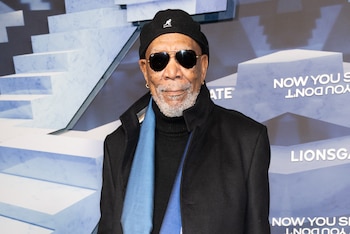 Morgan Freeman, con barba y bigote blancos, lleva gafas de sol, gorro negro, abrigo oscuro y bufanda azul, de pie frente a un fondo con texto y escalones