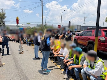 Grupo de personas sentadas en la acera junto a agentes de policía, con un SUV rojo y un cartel de McDonald's al fondo bajo un cielo azul claro