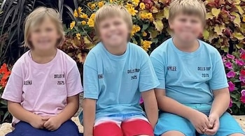 Tres niños de cabello rubio con las caras borrosas se sientan lado a lado, vistiendo camisetas de colores y pantalones cortos frente a un jardín floral.