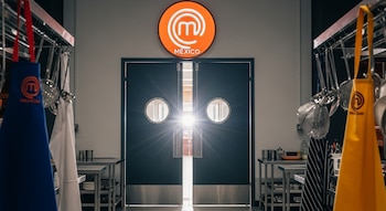 Vista interior de una cocina profesional de MasterChef México con puertas dobles abiertas y luz brillante, delantales azul y amarillo cuelgan en primer plano.