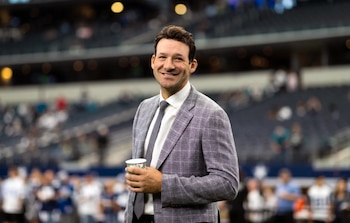 Hombre sonriendo, traje a cuadros gris, camisa blanca, corbata gris, vaso blanco. Fondo desenfocado de gradas de estadio con gente