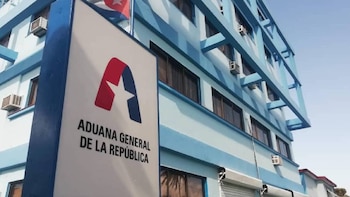 Vista exterior de un edificio azul y blanco, con un letrero que dice "Aduana General de la República" y el logo de una estrella blanca. Se ve una bandera cubana