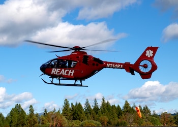 Un helicóptero rojo con la palabra REACH impresa en blanco vuela en un cielo azul con nubes sobre una arboleda