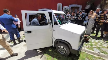 Un auto eléctrico blanco de diseño compacto con la puerta del conductor abierta, rodeado por personas que observan y toman fotografías en un evento