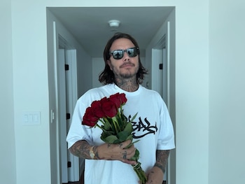 Sebastián Ortega, un hombre con cabello largo, barba y tatuajes, lleva gafas de sol y una camiseta blanca, sosteniendo un ramo de rosas rojas
