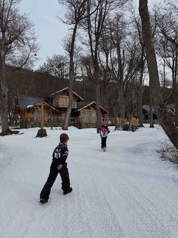 Dos niños de espaldas caminan sobre un sendero nevado. Al fondo, varias cabañas de madera con techos oscuros y árboles sin hojas