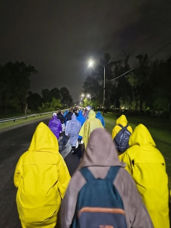 Personas de espalda vestidas con impermeables de colores caminan de noche por un camino asfaltado con farolas