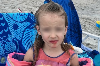 Una niña con manguitos flotadores y ojos difuminados sentada en una silla de playa junto a toallas azules sobre la arena.