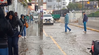 Peatones con abrigos caminan sobre una acera mojada y una calle con agua acumulada, con una camioneta blanca y edificios al fondo.