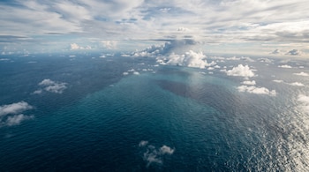 Vista aérea del océano con aguas de color azul y turquesa bajo un cielo con nubes blancas dispersas