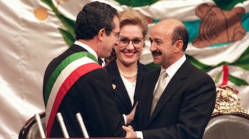 Dos hombres en traje formal se saludan de mano frente a una bandera mexicana, con una mujer sonriente al fondo.