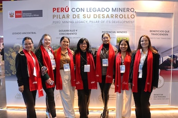 Siete mujeres vestidas con bufandas rojas y credenciales posan en fila delante de un panel publicitario del Ministerio de Energía y Minas