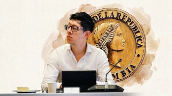 Un hombre joven con gafas y camisa blanca está sentado en una mesa con una tablet y un micrófono, con el emblema dorado de la República de Colombia de fondo.