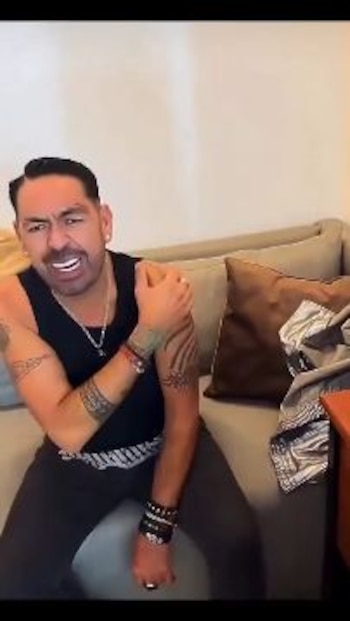 Un hombre con camiseta negra sin mangas y tatuajes se sienta en un sofá gris mientras apoya su mano derecha en el hombro izquierdo