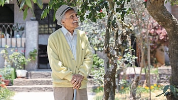Hombre mayor, con bigote y sombrero, vestido con camisa, cárdigan y pantalón, sostiene un bastón en un jardín con árboles y vegetación