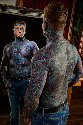 Un hombre con tatuajes en el rostro, la espalda y el torso se mira en un espejo que muestra su reflejo frontal