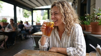 Mujer de cabello rubio rizado con camisa a rayas, sostiene un vaso de té helado con limón. Fondo borroso de un café con personas y plantas.