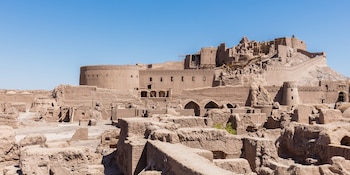 Gran ciudadela de adobe con altas murallas, varias torres y edificios de tierra, en medio de ruinas arquitectónicas y andamios bajo un cielo azul claro